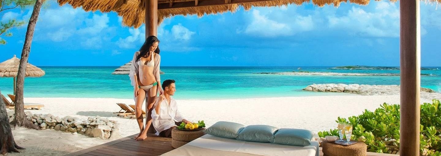 couple relaxing under a palapa by beach with cyrstal clear water in the background