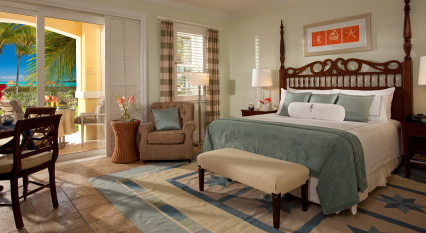 Beach House Oceanview Walkout Club Level Junior Suite- WOJ- Sandals Emerald Bay bed with sitting chair and a small table with the door to the balcony open at the beach house oceanview walkout club level junior suite at sandals emerald bay